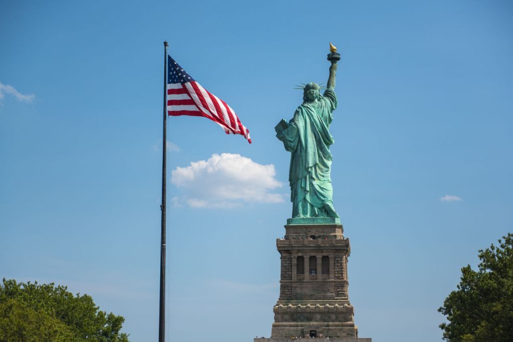 The Statue of Liberty alongside the American flag under a clear blue sky in New York City.