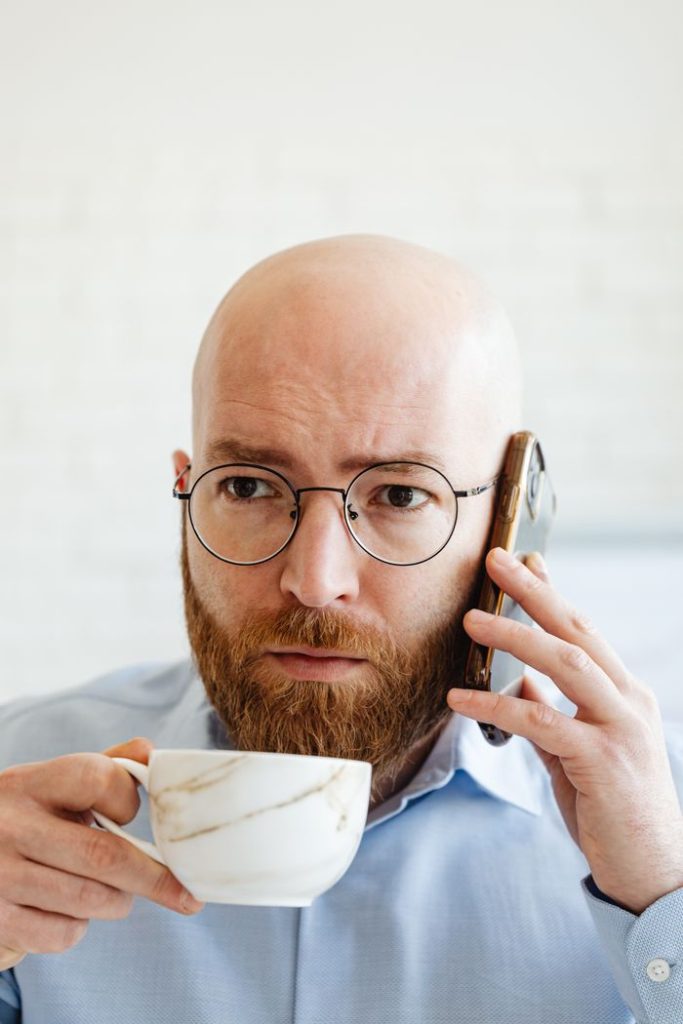Bald man with glasses using phone while drinking coffee in a bright setting.